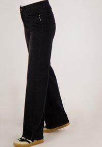 Zwarte high-waist rechte jeans gedragen met zwarte en beige sneakers, zichtbaar van midden taille tot voeten tegen een lichte achtergrond.