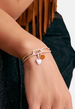 Handgelenk mit beigem Cord-Armband, das silberne Herz- und orangefarbene Edelsteinanhänger zeigt, getragen über dunklem Denim und brauner Kleidung mit Fransen.