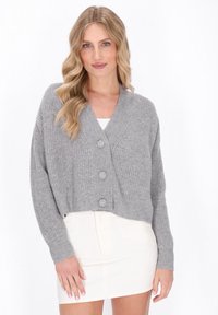 Grijze geribbelde cropped cardigan met een sjaalkraag, voorzien van drie grote knopen en lange mouwen, gecombineerd met een witte getailleerde jurk.