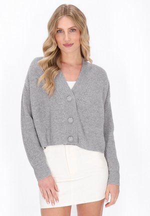 Cardigan corto grigio a coste con colletto a scialle, dotato di tre grandi bottoni e maniche lunghe, abbinato a un vestito bianco aderente.