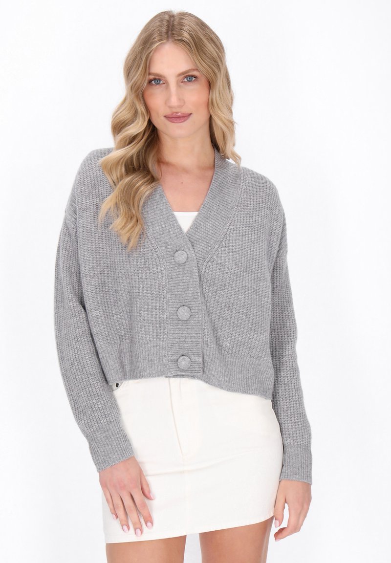 Grijze geribbelde cropped cardigan met een sjaalkraag, voorzien van drie grote knopen en lange mouwen, gecombineerd met een witte getailleerde jurk.
