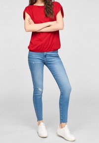 Top rosso a maniche corte con spalle arricciate, abbinato a jeans skinny strappati di un leggero blu. Sneakers bianche completano l'outfit.