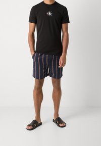 Svart bomullst-t-shirt med en logotyp, ihopparad med marinblå randiga shorts och svarta sandaler. Shortsen har vertikala ränder i röd och vit.