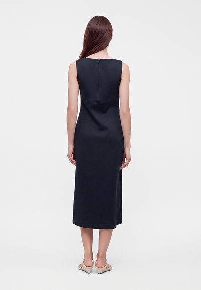 Femme aux longs cheveux bruns portant une robe midi noire sans manches vue de dos, debout sur un fond blanc avec des talons beiges.