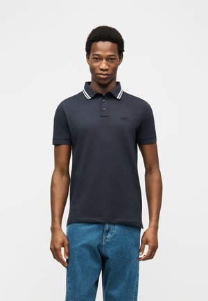 Άνδρας που φοράει μπλε πόλο μπλουζάκι με λευκή ριγέ γιακά και μπλε τζιν, στέκεται μπροστά σε απλό φωτεινό φόντο.