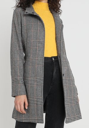 Grijs-zwart geruit mantel met een kleine kraag en lange mouwen, voorzien van een knoopsluiting en een comfortabele pasvorm. Draagt over een gele geribbelde trui.