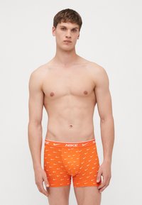Nike férfi narancssárga boxer alsónadrág, fehér swoosh mintázattal, elasztikus derékpánttal, testhezálló fazonnal és sima szövettextúrával.