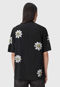 Osoba ubrana w czarną koszulkę z krótkim rękawem z białymi nadrukami stokrotek na plecach i rękawach, w połączeniu z ciemnymi jeansami.