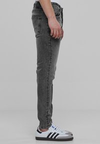 Personne portant un jean noir délavé slim et des baskets blanches Adidas avec des rayures noires, debout de profil sur un fond gris.