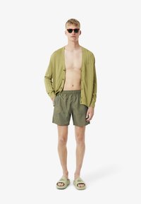 Olijfgroene geribde cardigan, gestreepte zwembroek in grijs met blauwe lijnen en groene sandalen. Model draagt een zonnebril en heeft blote voeten.