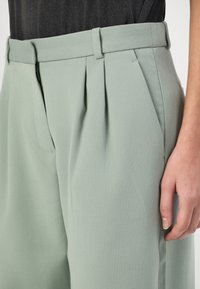 Mint green, pleated trousers with a smooth texture, featuring a zip closure and side pockets, displayed with a hand positioned near the waist.