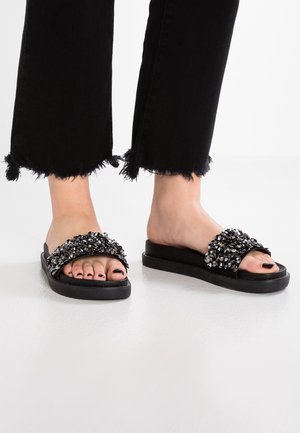 Musta liukusandaali, jossa leveä korukoristeltu hihna, jossa on kirkkaanvärisiä ja hopeisia koristeita, asetettuna mustalle teksturoidulle pohjalle.