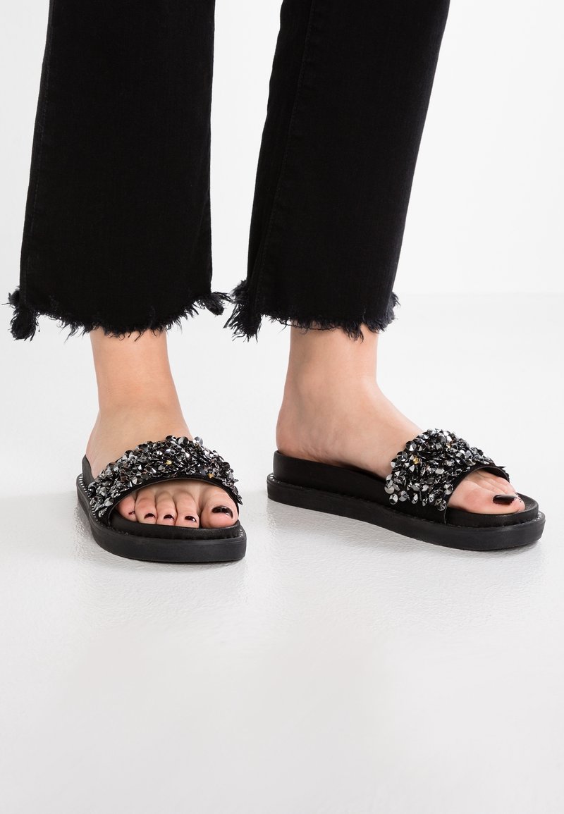 Sandales à glissière noires avec une large bride ornée de bijoux comprenant des embellissements clairs et argentés, posées sur une semelle noire texturée.