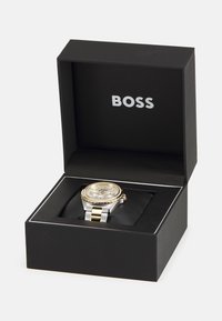 Orologio argento e oro in scatola nera, con quadrante rotondo e dettagli dorati, bracciale in metallo e il nome del marchio "BOSS" sulla cima.