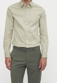 Uomo che indossa una camicia button-up beige chiara infilata in pantaloni tailored verde oliva con passanti per la cintura, in piedi di fronte a uno sfondo semplice.