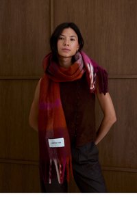 Oversized sjaal in gradient tinten van rood en roze met franjes en een label dat "ARCTIC FOX" leest, gecombineerd met een bruin mouwloos topje.