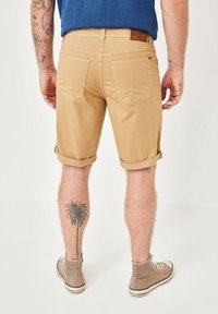 Tan cotton shorts with rolled hems, featuring two back pockets and a brown leather patch on the waistband. A palm tree tattoo is visible on the leg.