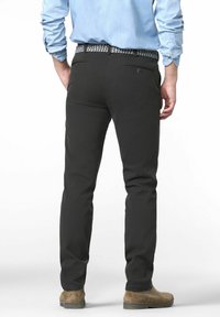 Mann in schwarzen Hosen, hellblauem Langarmhemd, geflochtenem Gürtel und braunen Wildleder-Schuhen, der mit dem Rücken zu einer weißen Hintergrund steht.