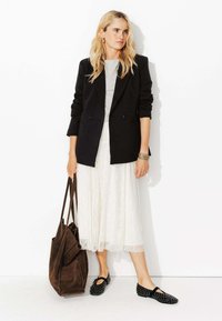 Blazer noir croisé, t-shirt gris, jupe blanche en dentelle, sac fourre-tout en suède marron, ballerines noires avec découpes rondes. Look de mode minimaliste.