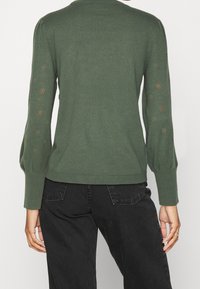 Pull en tricot vert avec des manches longues, présentant des détails bouffants et un motif en dentelle le long des poignets. Ourlet côtelé à la taille.