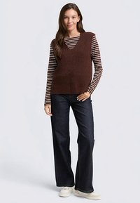 Gilet marrone lavorato a maglia con scollo a V sopra una camicia a righe a maniche lunghe, abbinato a jeans scuri a gamba larga e scarpe color crema.