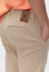 Pantaloni beige con una consistenza liscia, dotati di un'etichetta in pelle marrone con logo impresso e un accento in cordoncino verde lime vicino alla vita.