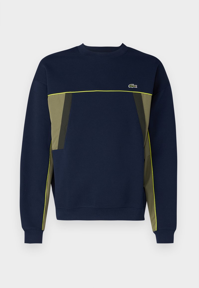Sweatshirt bleu marine avec des accents de panneaux verts et gris, présentant un col rond et des passepoils jaunes. Logo Lacoste sur le haut du côté gauche de la poitrine.