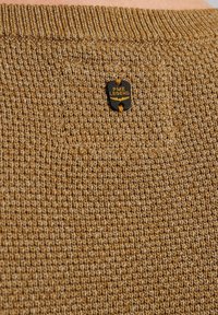 Sweter z dzianiny w ciepłym brązowym kolorze, z teksturowanym wzorem oraz metką w kolorze czarnym i złotym z logo PME Legend blisko dekoltu.