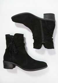 Svarta mocka ankelboots med låg, staplad klack, sidsdragkedja och dekorativa fransar vid ankeln. Strukturerad yta med vågiga kanter.