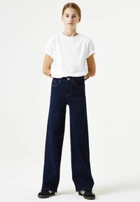 Wijd uitlopende donkere denim jeans met een hoge taille, gecombineerd met een aansluitend wit katoenen T-shirt. Model staat tegen een effen achtergrond.