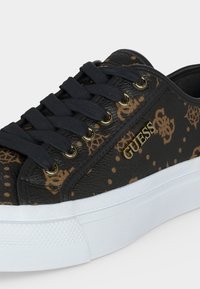 Μαύρο και καφέ αθλητικό παπούτσι Guess με μαύρα κορδόνια, χρυσά ανοίγματα, μεταλλικό λογότυπο Guess και παχύ λευκό καουτσούκ σόλα.