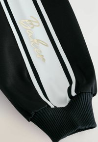 Čierna športová sako s rukávom, ktorý má biely pruh a zlaté nápisy "Baker". Textúrovaná tkanina s rebrovanými manžetami.