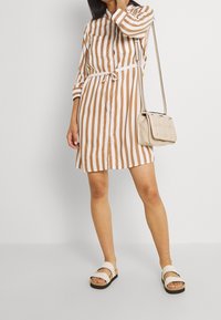 Donna che indossa un vestito a righe verticali marroni e bianche con bottoni e vita con lacci, sandali beige e una borsa a spalla beige, su uno sfondo bianco.