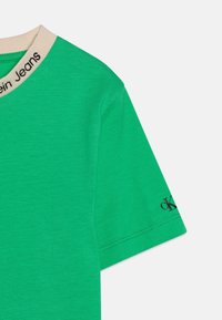 Grön bomullströja med rund hals, beige krage och svart "ck"-logotyp på vänster ärm. Mjuk textur och korta ärmar.