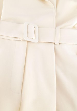 Crèmekleurige stof met bijpassende riem en rechthoekige gesp, met zichtbare plooien en stikseldetails.