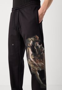 Black sweatpants featuring an artistic print on the left leg, elastic waistband, and side pockets. Smooth texture and relaxed fit.