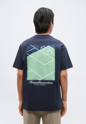 Άνδρας που φοράει μπλουζάκι ναυτικού χρώματος με μινιμαλιστικό γραφικό γηπέδου ποδοσφαίρου και το κείμενο "Scandinavian Football Community" στην πλάτη.