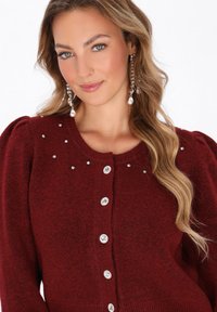Cardigan a maglia borgogna con scollo rotondo, impreziosito da strass, presenta maniche a palloncino e sei bottoni decorativi.