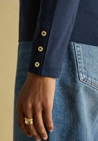 Sinine pikkade varrukatega pluus kolme kuldse nööbiga mansetil, paaritatud kõrge vöökohtadega siniste teksadega. Sile kangakonstruktsioon on selgelt nähtav.