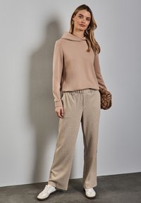 Felpa beige con cappuccio abbinata a pantaloni a gamba larga con fantasia a quadri. Il modello tiene una borsa intrecciata marrone; le sneakers bianche completano l'outfit.