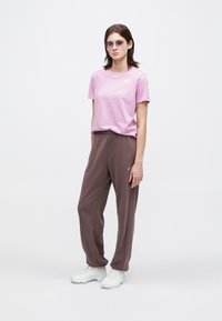 Νέος άνθρωπος φοράει μωβ γυαλιά ηλίου, ροζ μπλούζα Nike, καφέ παντελόνι Nike και λευκά αθλητικά παπούτσια, στέκεται σε λευκό φόντο.