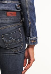 Gros plan d’une personne portant un jean en denim bleu et une veste, montrant la poche arrière avec une étiquette en cuir LTB et le bouton sur le poignet de la veste.