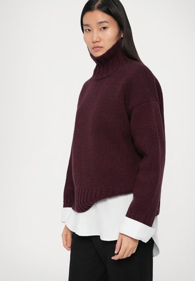 Pull en mailles bourgogne à col roulé avec ourlet côtelé, porté par-dessus une chemise blanche à col avec texture côtelée, associé à un pantalon noir.
