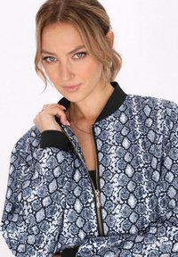 Jachetă bomber cu un model de imprimeu cu șarpe în nuanțe de navy și alb, dotată cu un guler ribetat negru, fermoar în față și o textură de țesătură moale.