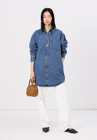 Denim srajčna obleka v srednji modri barvi, zaključen s gumbi, s prsno žepom, sproščen kroj, ukrivljen rob, oblečena preko belih hlač. Rjav nahrbtnik in usnjeni čevlji s koničastimi prsti.