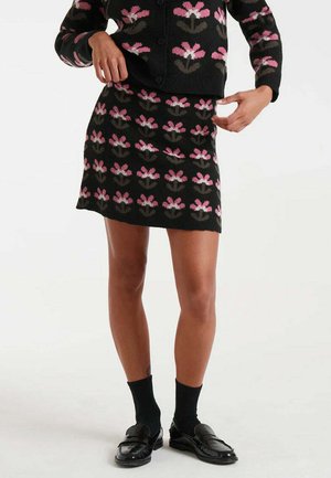 Falda de punto negra con un patrón floral en rosa y gris. Combinada con un cárdigan a juego, usada con calcetines negros y mocasines.