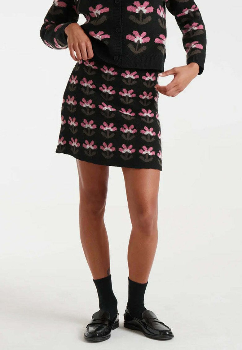 Falda de punto negra con un patrón floral en rosa y gris. Combinada con un cárdigan a juego, usada con calcetines negros y mocasines.