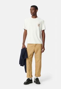 Wit katoenen t-shirt met "LON PRS" in het bruin op de borst gedrukt, gecombineerd met kaki broek en zwarte schoenen, en een marineblauwe trui vasthoudend.