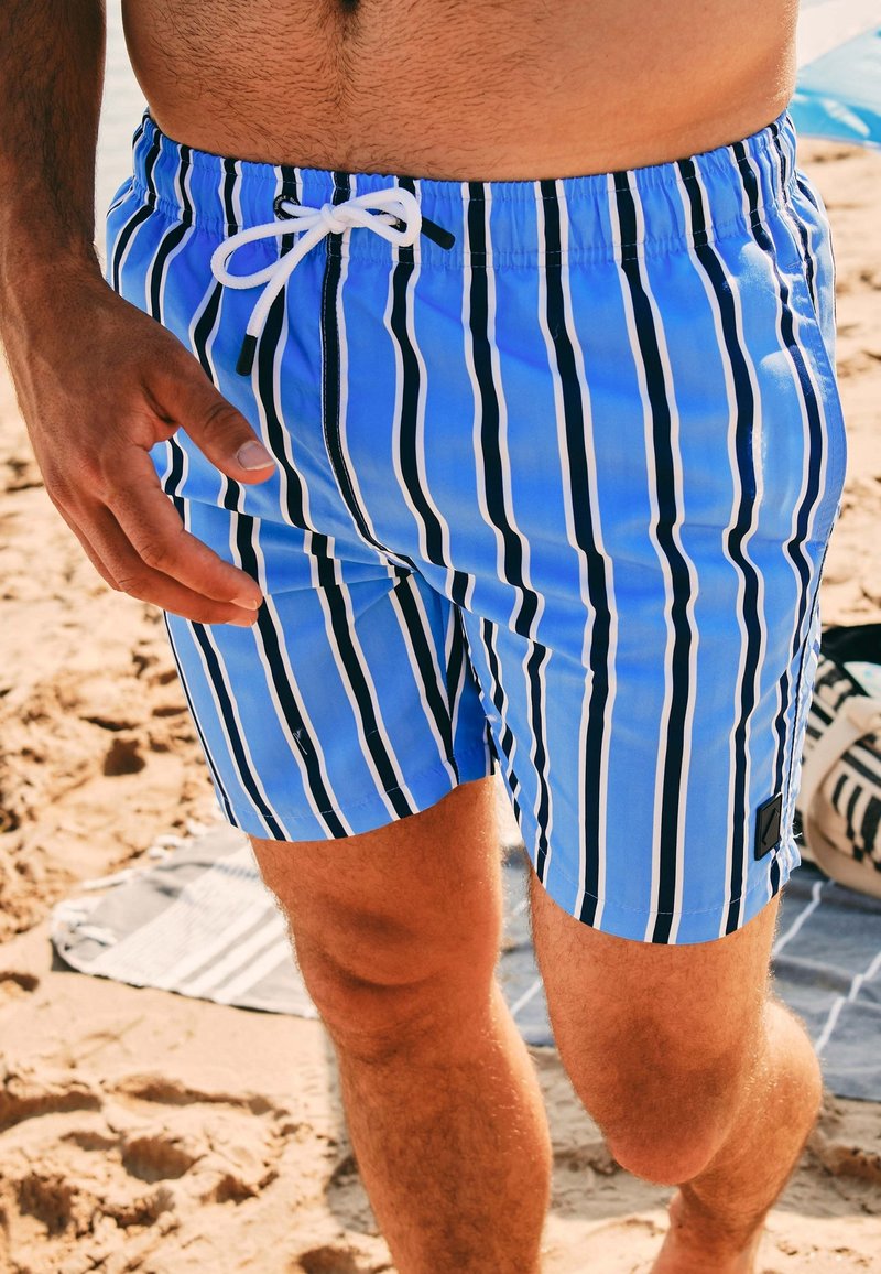 Blauwe gestreepte zwembroek met witte en zwarte strepen, elastische tailleband, witte trekkoord en een logo-patch aan de rechteronderkant.
