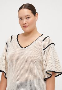 Cream-colored mesh top with a V-neck, black scalloped trim, and short, flared sleeves; features an open weave pattern and relaxed fit.
